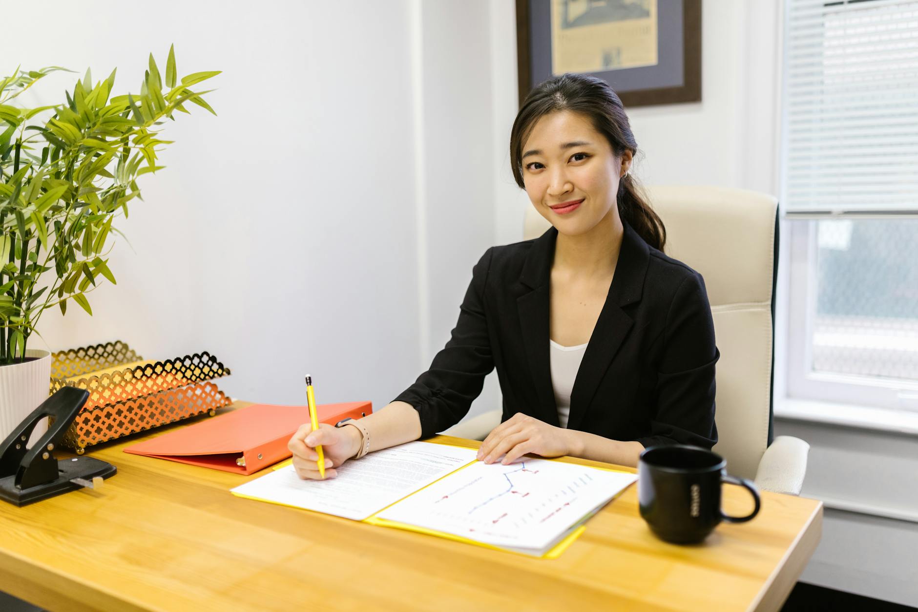 Businesswoman at desk writing with a pen, smiling confidently in a modern office